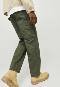 Pantaloni cargo de culoare verde măsliniu, cu mai multe buzunare, croi lejer și material ușor, asortați cu bocanci bej și o cămașă ușoară.