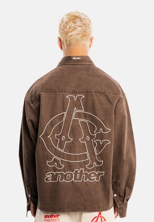 Persona con cabello rubio corto que lleva una chaqueta marrón con grandes letras cosidas y la palabra "another" en la espalda.