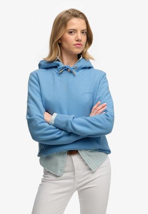 Jeune femme aux cheveux blonds portant un sweat à capuche bleu par-dessus une chemise rayée et un pantalon blanc, debout bras croisés devant un fond blanc.