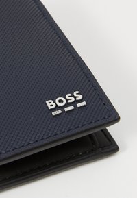 Tumši jūras zilas teksturētas maks, ar sudraba izsistā "BOSS" logo, kas piedāvā kontrastējošas šuves un slaidu, taisnstūrveida formu.