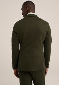Chaqueta de traje verde oscuro, entallada, con una textura suave, cuello con muescas y dos aberturas traseras. Incluye puños abotonados y hombros naturales.