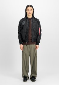 Chaqueta bomber negra con detalles acolchados, cierre frontal y parches. Usada sobre una camiseta marrón, combinada con pantalones holgados verde oliva y zapatos negros.