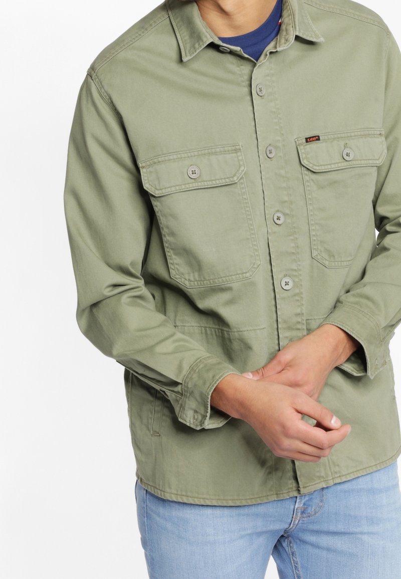 Homme portant une chemise légère vert olive à boutons avec poches poitrine et un jean bleu clair, mains jointes à la taille.