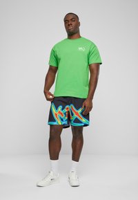 Grønn bomull t-skjorte med korte ermer, kombinert med svarte shorts med et fargerikt, bølgete mønster. Hvite sneakers og ankelsokker fullfører looken.
