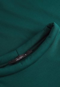 Material verde cu textură netedă; prezintă un guler rotund și un etichetă etichetată "Jascha" cu mărimea "S" near marginea pliată.