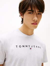 Graues Baumwoll-T-Shirt mit dem "TOMMY JEANS" Logo in schwarzem Stick und einem kleinen rot-weißen-blauen Flaggenakzent auf der linken Seite.