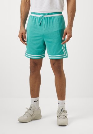 Homme portant un short de basket bleu sarcelle avec bordure blanche, des chaussettes de sport blanches et des chaussures de sport beiges, debout devant un fond blanc.