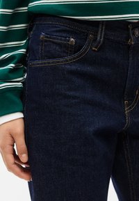 Κοντινό ενός ατόμου που φοράει σκούρο μπλε τζιν και μακρυμάνικο πουκάμισο με πράσινες και λευκές ρίγες, με το χέρι να ακουμπάει στο πλάι.