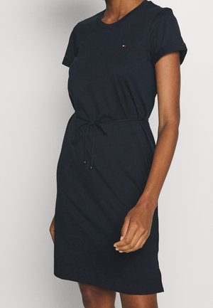 Femme portant une robe noire à manches courtes avec une ceinture nouée à la taille et un petit logo sur la poitrine et la manche.