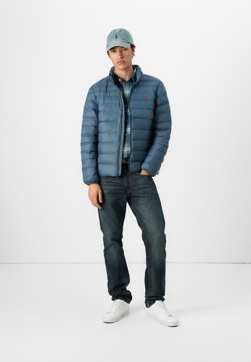 Chaqueta de plumas azul con secciones acolchadas horizontales, combinada con una camisa de cuadros de botones, jeans oscuros y zapatillas blancas; lleva una gorra azul claro.
