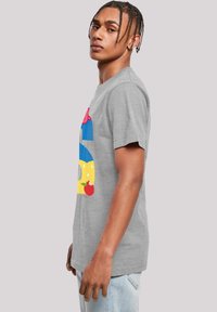 Pelēka kokvilnas t-krekls ar krāsainu grafisko dizainu, kurā redzama zila cepure un dzeltenas detaļas, prezentēts vīriešu modelī no sāna.