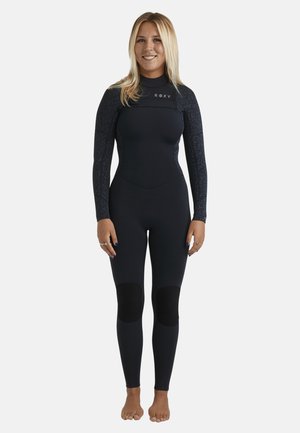 Kobieta o blond włosach ubrana w czarny piankowy kombinezon z wzorzystymi rękawami i ochraniaczami na kolanach, stojąca boso na białym tle.