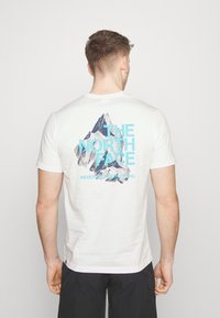 White cotton t-shirt featuring a mountain graphic in blue and dark gray on the back, with "THE NORTH FACE" and "NEVER STOP EXPLORING" text.