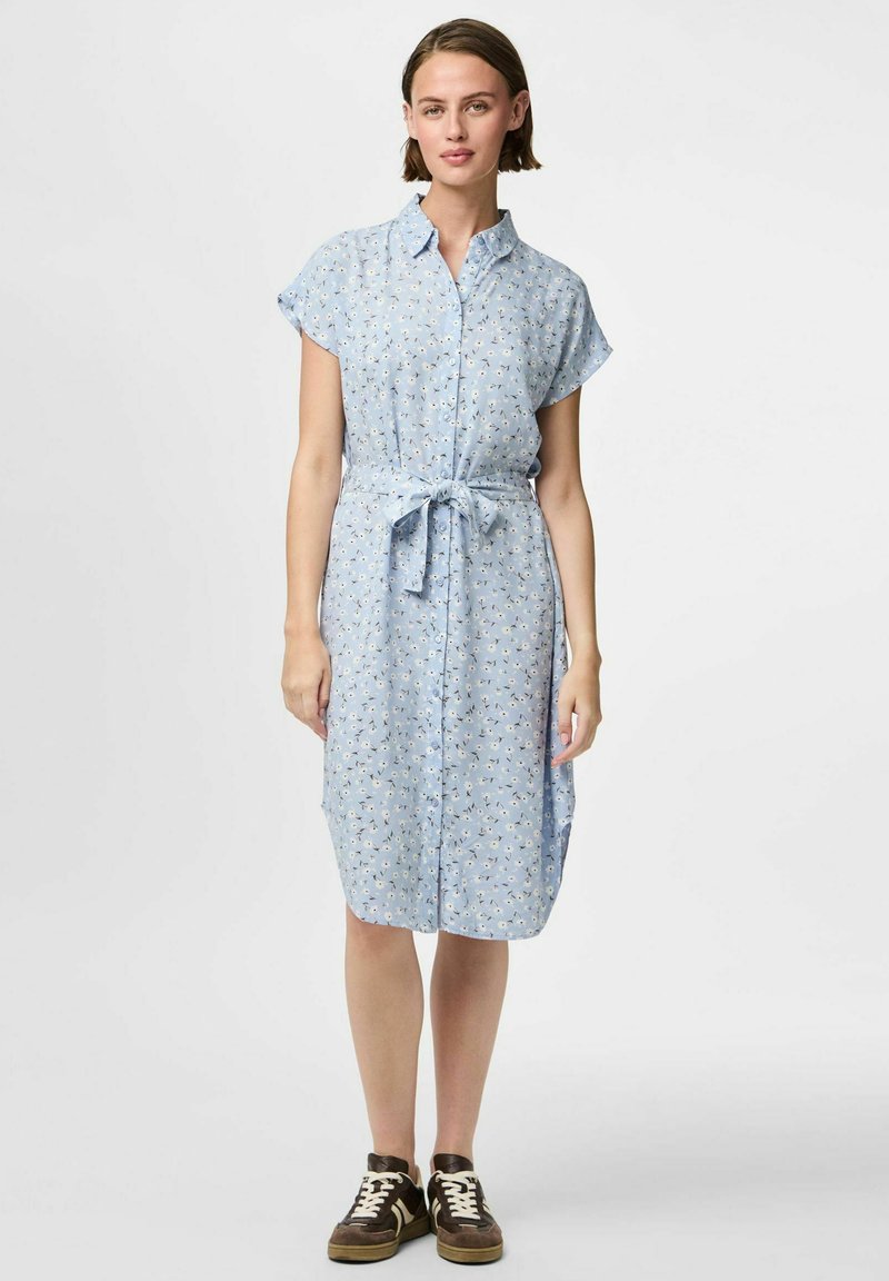 Ljusblå, knappad klänning med korta ärmar, bälte i midjan, blommönster och knälång fåll. Styckad med bruna sneakers.