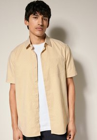 Camisa amarilla de manga corta con un patrón geométrico, que cuenta con un diseño de botones y un bolsillo frontal sobre una camiseta blanca.