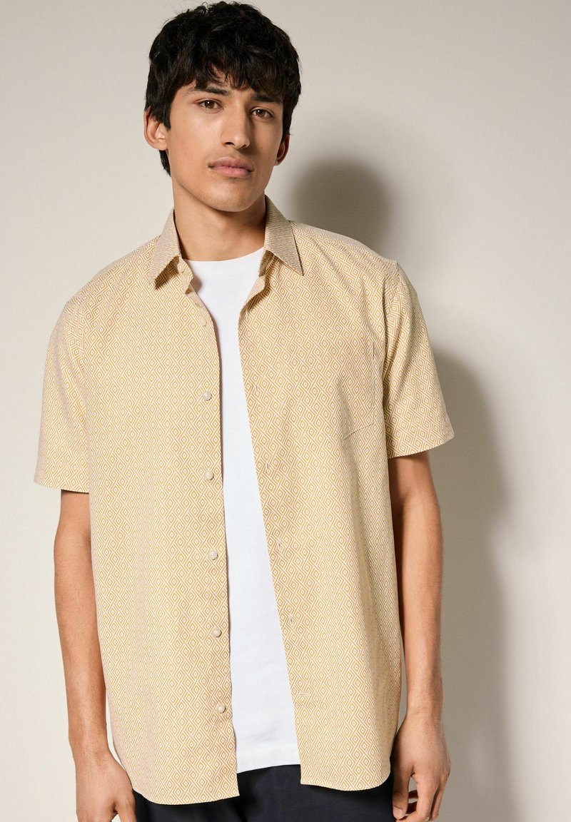 Camisa amarilla de manga corta con un patrón geométrico, que cuenta con un diseño de botones y un bolsillo frontal sobre una camiseta blanca.