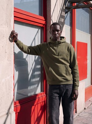 Jeune homme en sweat à capuche vert et jean foncé appuyé contre une porte en verre à cadre rouge par une journée ensoleillée avec des ombres sur le mur.