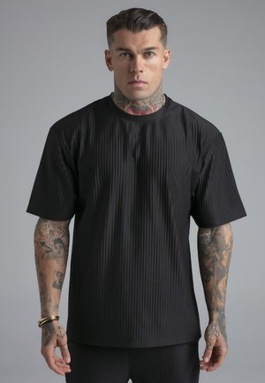 Mann mit Tätowierungen am Hals und an den Armen, der ein schwarzes, geripptes Kurzarmshirt trägt und vor einem einfarbigen grauen Hintergrund steht.