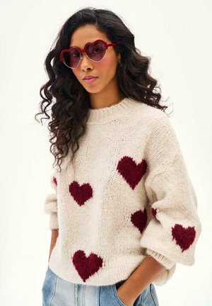 Kobieta z długimi kręconymi włosami, nosząca czerwone okulary przeciwsłoneczne w kształcie serca oraz biały sweter ozdobiony czerwonymi sercami, z rękami w kieszeniach jeansów.