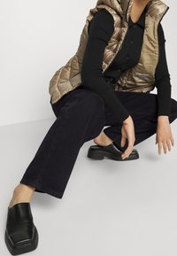 Persona en cuclillas con camisa negra abotonada, chaleco acolchado dorado, pantalones anchos negros y zapatos sin cordones negros frente a un fondo blanco.