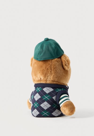 Brauner Teddybär trägt grüne Mütze und dunkelblaue Argyle-Strickweste mit gestreiften Ärmeln, von hinten auf einfachem Hintergrund gezeigt.