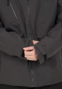 Donkere waterdichte jas met een gladde textuur, voorzien van contrasterende ritszakken, verstelbare manchetten en een veilige ritssluiting aan de voorkant.