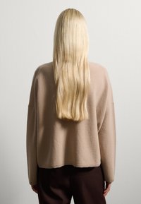 Maglione di lana beige con vestibilità morbida, spalle basse e orlo a coste, abbinato a pantaloni marroni scuri. Capelli lunghi, lisci e con riflessi visibili.