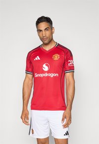 Červený dres Manchester United s čiernymi akcentmi, v-krčný dizajn, biele šortky, s logami Snapdragon a Adidas na prednej časti a rukávoch.