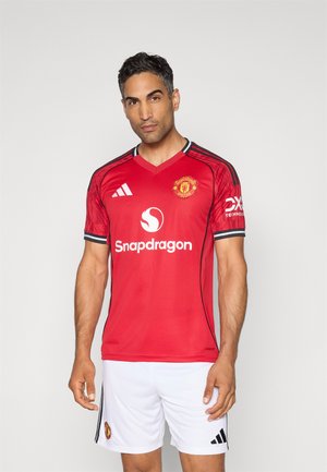 Maillot rouge de Manchester United avec des accents noirs, design en v, short blanc, arborant les logos de Snapdragon et Adidas sur le devant et les manches.