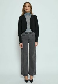 Schwarze offene Strickjacke, grauer Rollkragenpullover, hochgeschnittene weiße Jeans mit weitem Bein, schwarze spitze Absätze. Glattes Material und maßgeschneiderte Passform.