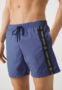 Pantaloni da bagno blu con cintura elastica e cordoncino. Presenta un pannello laterale nero con la scritta "TOMMY JEANS" in bianco. Texture liscia.