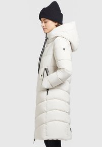 Witte, puffy jas met een capuchon, voorzien van rits-zakjes aan de zijkanten, zwarte trekkoorden en een chevron stikpatroon, gedragen met een zwarte beanie.