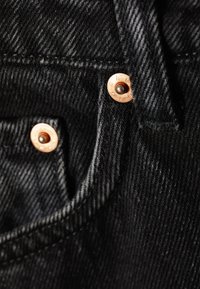 Nahaufnahme von schwarzem Denimstoff mit Kupfernieten und sichtbaren Nähten an einer Taschenkante.