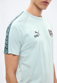 Homme portant un maillot de sport bleu clair avec l'inscription "KING" et le logo Puma, ainsi qu'un écusson de football d'Autriche sur la poitrine, avec une bande à motifs sur l'épaule latérale.