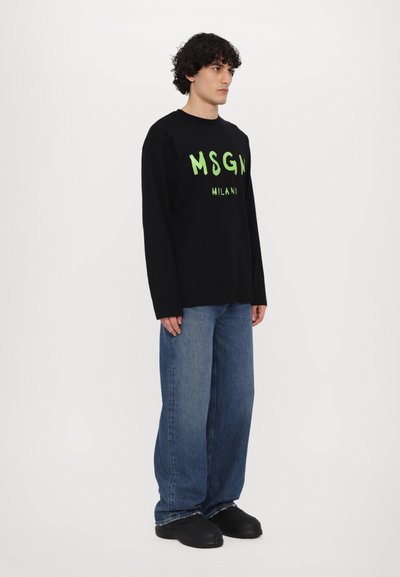 Svart langermet skjorte med grønn "MSGM MILANO" trykk, parret med løse blå jeans og svarte sko, som viser en avslappet passform og uformell stil.