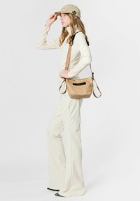 Bolso bandolera beige con cuero texturizado, correa ajustable y detalle de etiqueta negra. Modelo con conjunto crema y gorra beige.