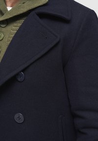 Tumši zils jūras stila mētelis ar lielām pogām pār olīvu zaļa džempera apkakli ar līdzīgām pogām.