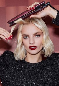 GHD Haarglätter in Burgunderrot mit edlem metallischen Finish, in der Nähe von glänzendem blonden Haar gehalten. Paillettenoberteil in Schwarz und funkelnde Ohrringe sind sichtbar.