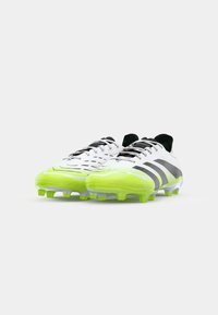 Des chaussures de football blanches et noires avec des accents jaunes. Elles possèdent une tige texturée et des pointes en caoutchouc pour une meilleure traction. Les lacets sont placés au centre.