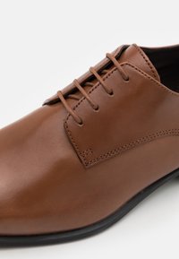 Scarpa in pelle marrone con una finitura liscia, caratterizzata da un design con lacci chiusi, dettagli di cucitura sottili e una suola nera.