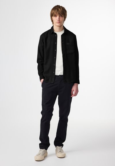 Camicia nera con bottoni sopra una maglietta bianca a girocollo, abbinata a pantaloni scuri e scarpe da ginnastica beige. La camicia presenta una tasca sul petto e un taglio morbido.