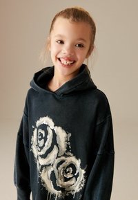 Haut à capuche noir avec un grand graphique de rose blanche sur le devant. Texture douce, coupe oversize et capuche avec cordon de serrage.
