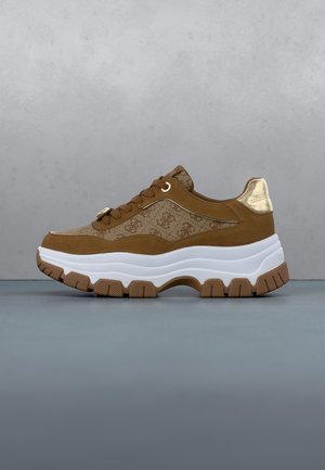 Brown and beige sneaker with chunky white sole, patterned fabric sides, brown laces, and metallic gold heel accent on a gray surface.