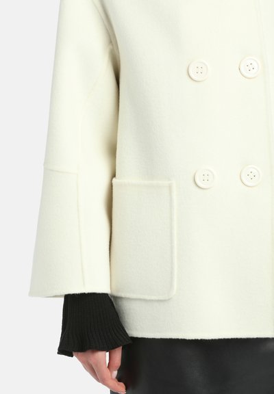 Kaksirivinen kerma-valkoinen villakangastakki suurilla valkoisilla nappeilla, sivutasku ja teksturoitu musta neulottu hihansuu. Tasainen pinta kauttaaltaan.