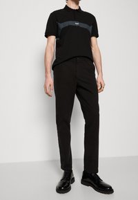 Černé polo tričko se šedým pruhem, logo na hrudi, v kombinaci s černými slim-fit kalhotami a lesklými černými šněrovacími botami.