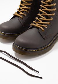 Bottes en cuir marron à lacets avec des coutures jaunes et des bouts ronds, présentées avec un lacet desserré sur une surface blanche.