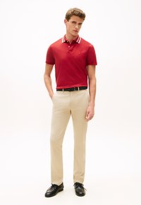 Κόκκινο πόλο με γιακά που έχει λευκές ρίγες, συνδυασμένο με μπεζ παντελόνι και μαύρα παπούτσια. Το ντύσιμο έχει εφαρμοστή γραμμή και ομαλή υφή.