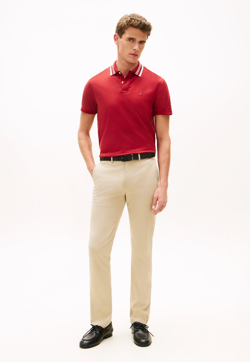 Κόκκινο πόλο με γιακά που έχει λευκές ρίγες, συνδυασμένο με μπεζ παντελόνι και μαύρα παπούτσια. Το ντύσιμο έχει εφαρμοστή γραμμή και ομαλή υφή.