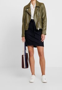 Veste de motard en cuir olive avec des accessoires en argent, associée à une jupe en denim foncé. Des baskets blanches et un sac color block complètent le look.