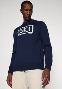 Námořnický modrý svetr z jemné pleteniny s bílým logem "SKI" na hrudi a žebrovanými manžetami a spodním lemem.
